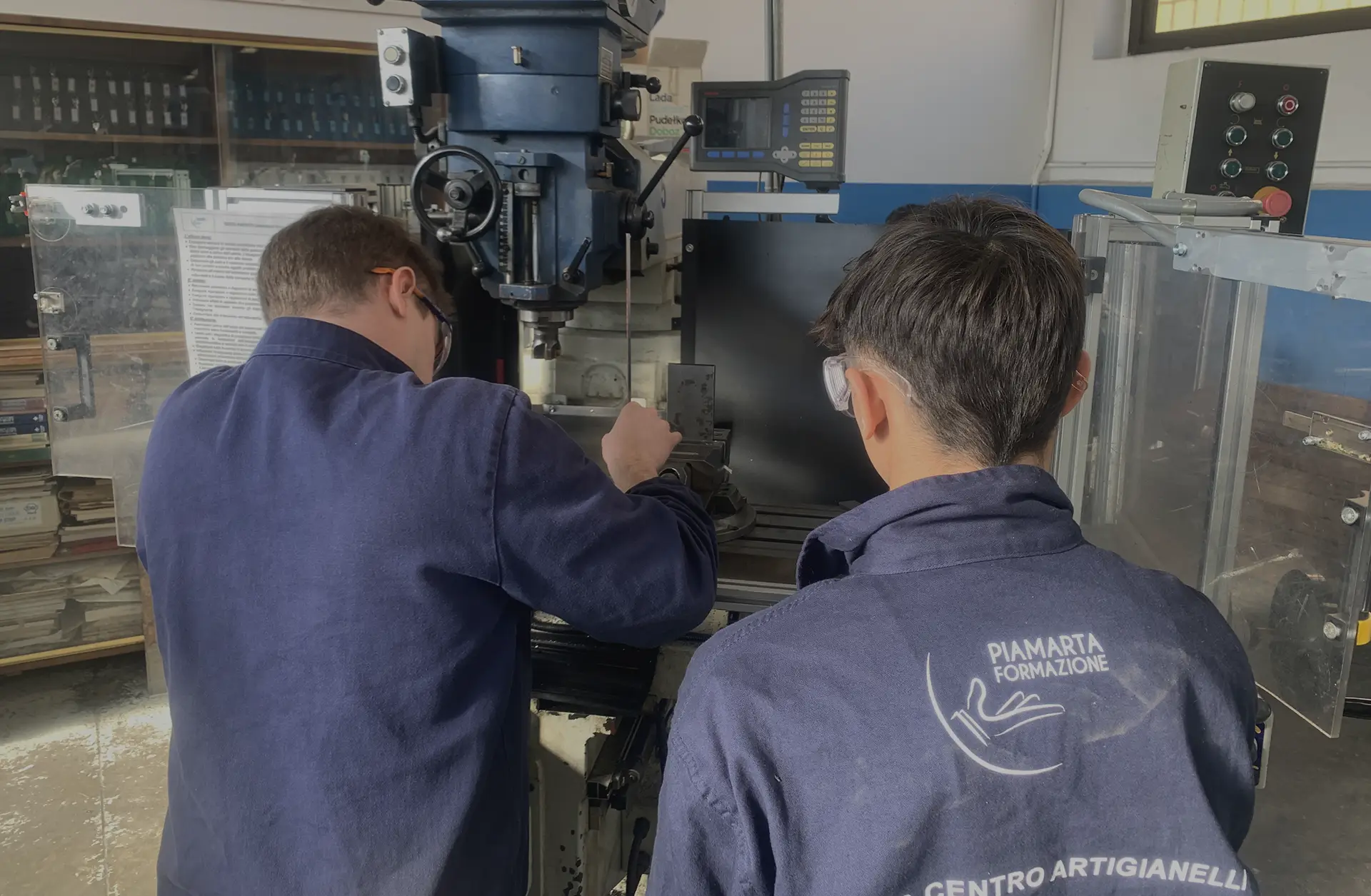 Due studenti lavorano su una macchina utensile in un'officina, indossando camici da lavoro e occhiali protettivi.
