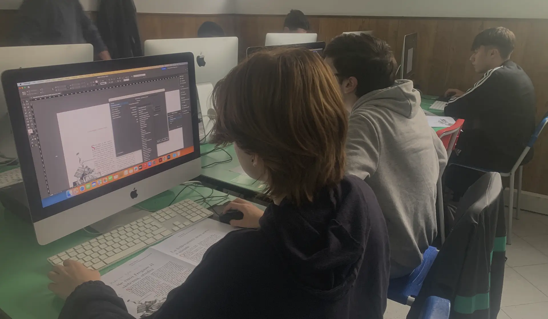 Studenti al computer durante una lezione di grafica, lavorano su un progetto utilizzando software di impaginazione su iMac. Sullo schermo è visibile un documento aperto con testo e immagini.