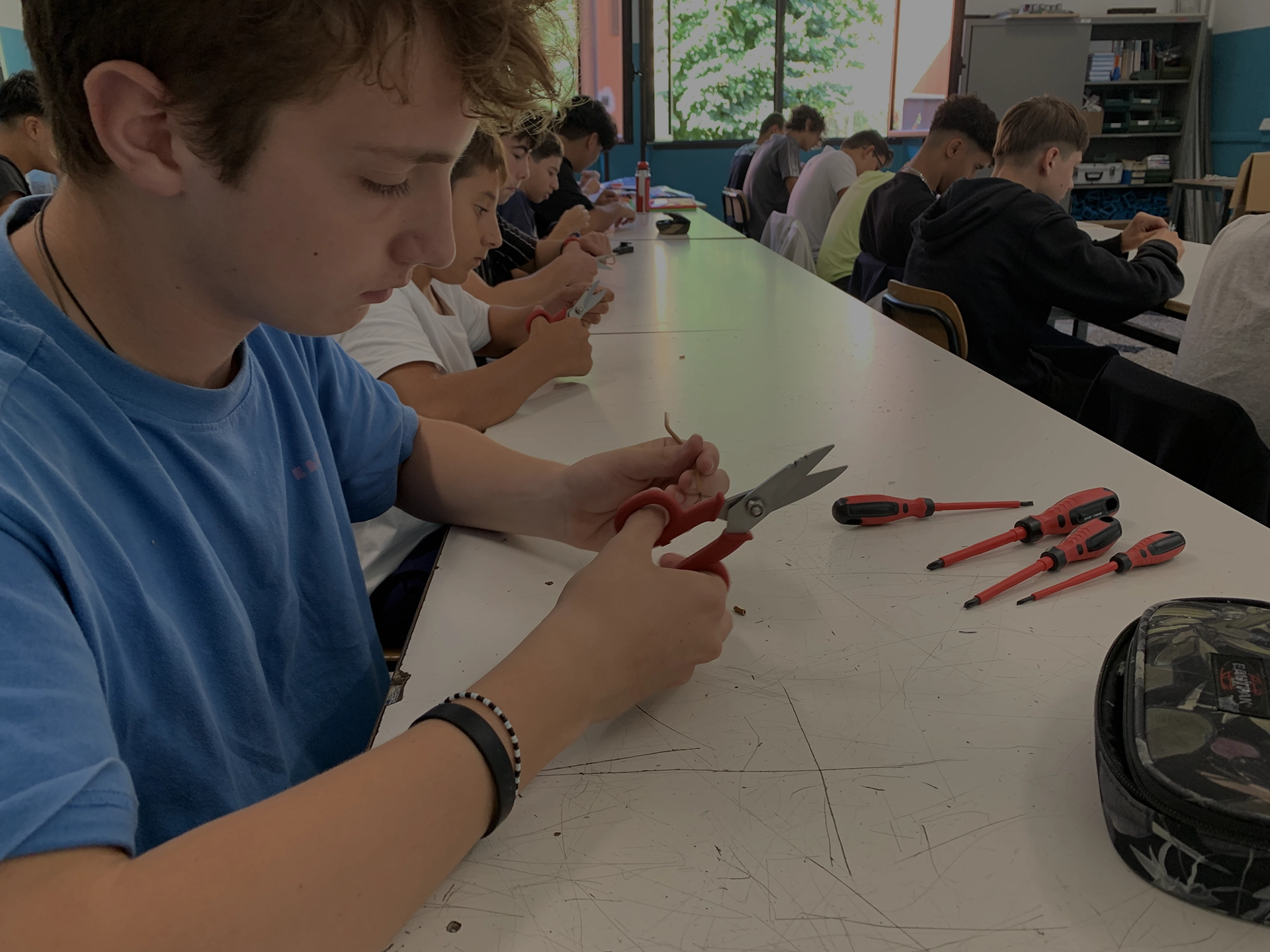 Studente che utilizza forbici per tagliare un cavo, circondato da cacciaviti su un tavolo di lavoro.