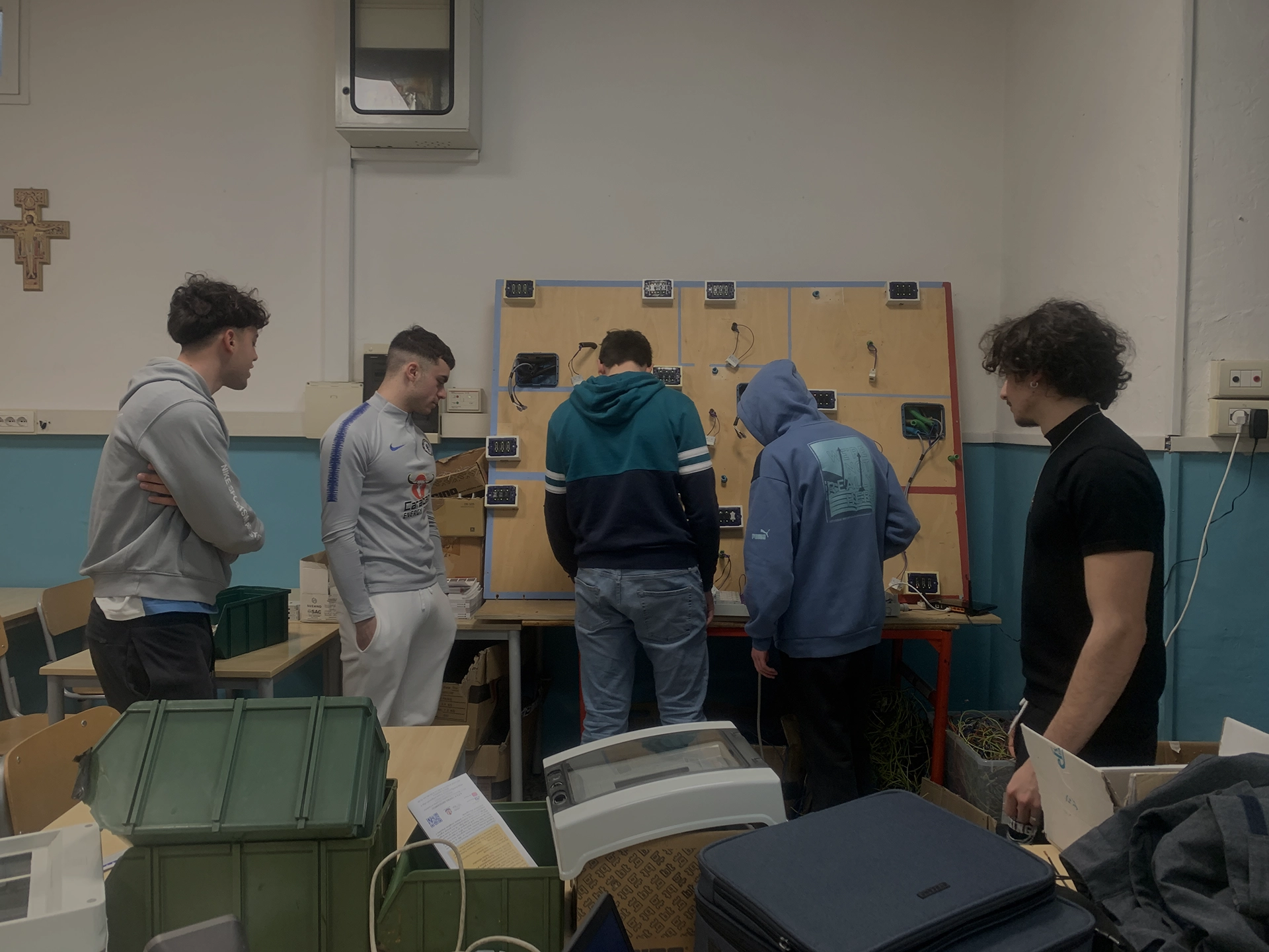 Cinque ragazzi in un laboratorio osservano e lavorano su una parete con collegamenti elettrici.