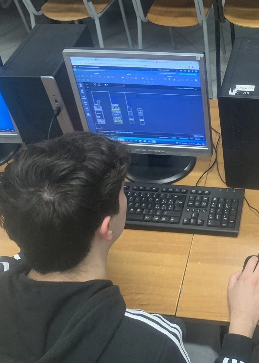 Un gruppo di studenti è concentrato davanti ai computer in un'aula informatica, mentre uno di loro offre supporto al compagno, guardando insieme lo schermo.