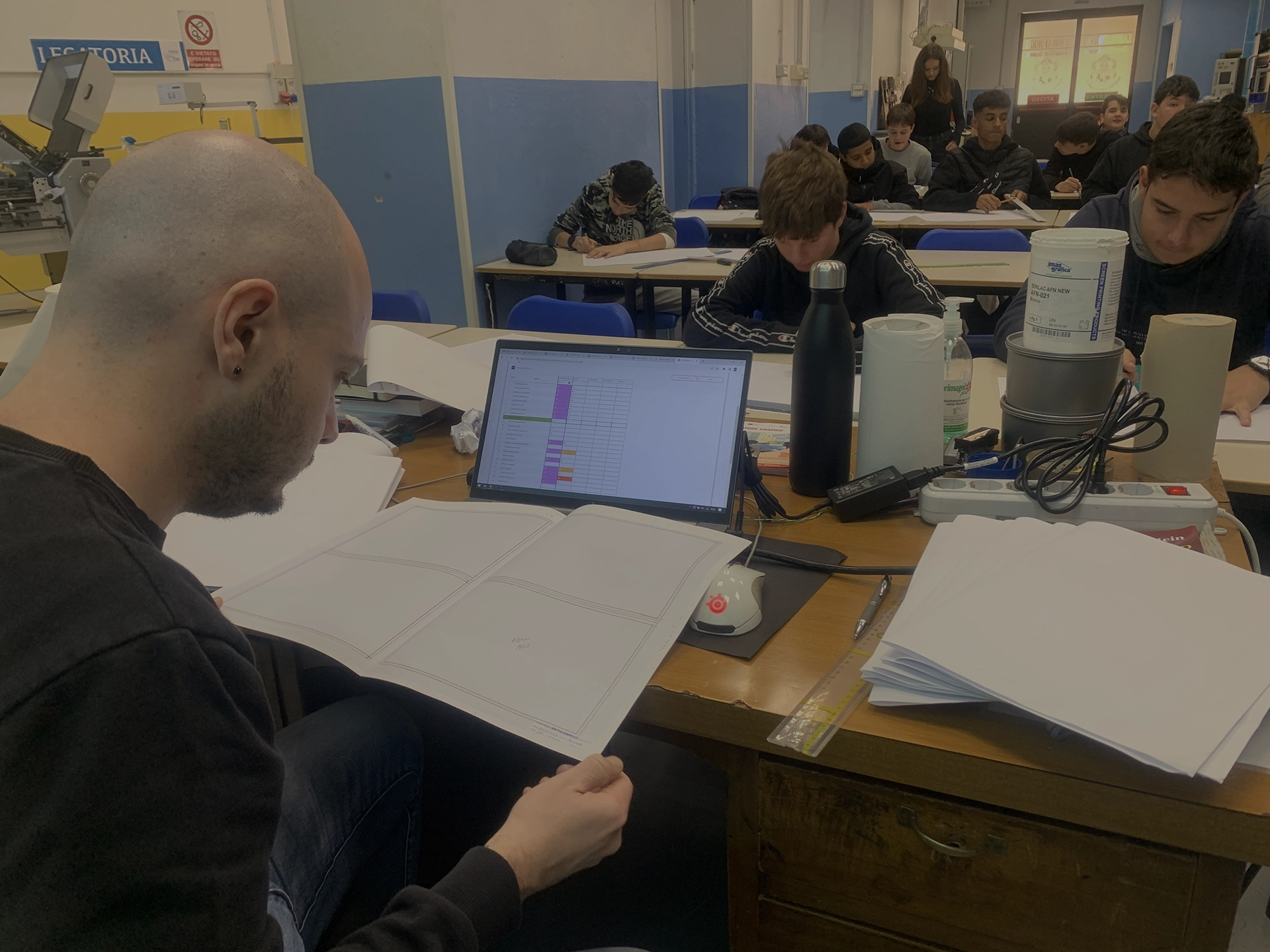 Un insegnante sta correggendo dei documenti su un tavolo con un computer portatile aperto di fronte a lui, mentre gli studenti in classe sono concentrati sul lavoro.