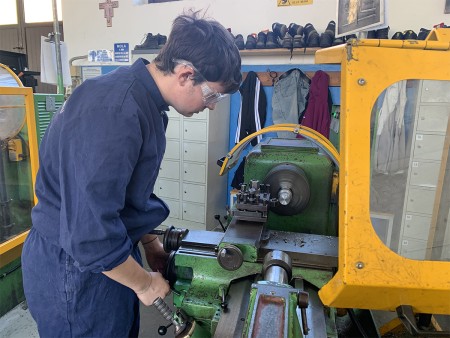 Studente utilizza un tornio meccanico indossando occhiali protettivi e tuta da lavoro, eseguendo una lavorazione di precisione su un pezzo di metallo.