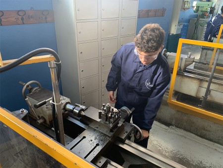 Studente che utilizza un tornio meccanico per una lavorazione di precisione in officina, indossando occhiali protettivi e una divisa da lavoro.