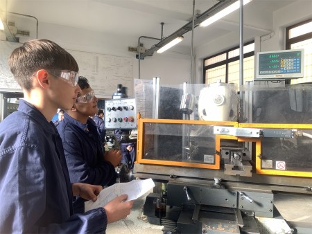 Due studenti con occhiali di sicurezza osservano una fresatrice, uno dei quali tiene in mano un disegno tecnico.