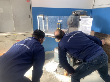 Due persone in tuta da lavoro osservano una macchina utensile protetta da una copertura trasparente, all'interno di un laboratorio tecnico.