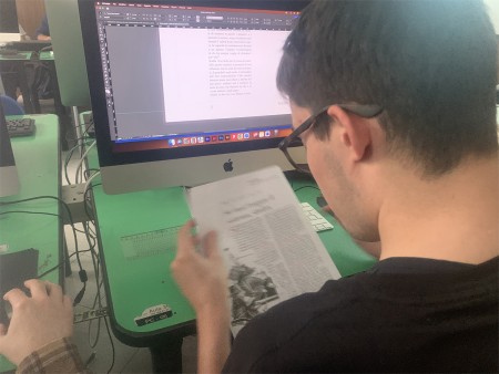 Studente consulta un documento cartaceo mentre lavora su un progetto di grafica al computer.