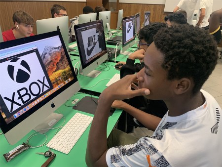 Studente che osserva un progetto di grafica su un computer, con i loghi Xbox e Nike visualizzati sugli schermi.
