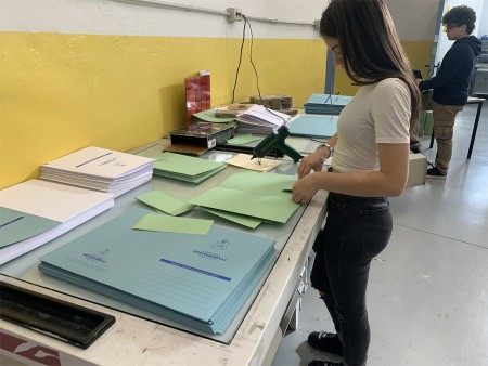 Studentessa prepara fascicoli piegando fogli di carta su un tavolo in laboratorio.