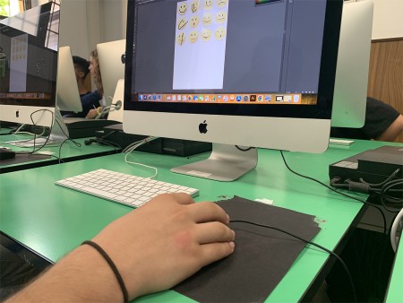 Primo piano della mano di uno studente mentre utilizza il mouse, lavorando su un progetto grafico con emoji su un computer in aula informatica.