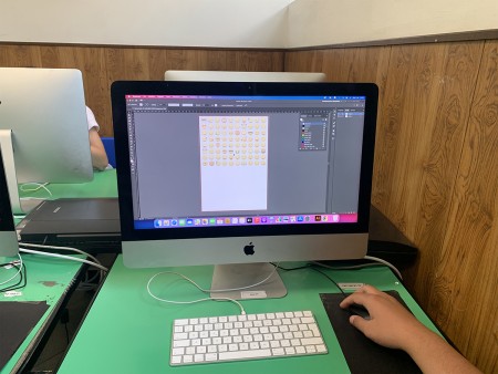 Primo piano di un monitor su cui è aperto un programma di grafica con vari emoji, mentre una mano utilizza il mouse. L'aula informatica è visibile sullo sfondo.