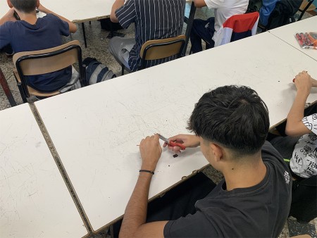 Studente al lavoro con un attrezzo manuale su un tavolo graffiato.