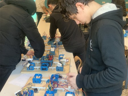 Studenti impegnati in un'attività pratica di cablaggio elettrico in classe, mentre collegano componenti su pannelli di legno con scatole elettriche blu e fili colorati.