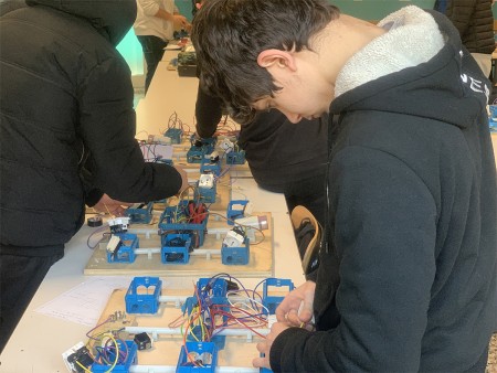  Studente che lavora su un progetto di cablaggio elettrico, collegando fili su una tavola con scatole elettriche blu, mentre altri studenti eseguono attività simili sullo sfondo.