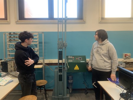 Due studenti osservano un macchinario industriale in un laboratorio scolastico, probabilmente discutendo del suo funzionamento o utilizzo.