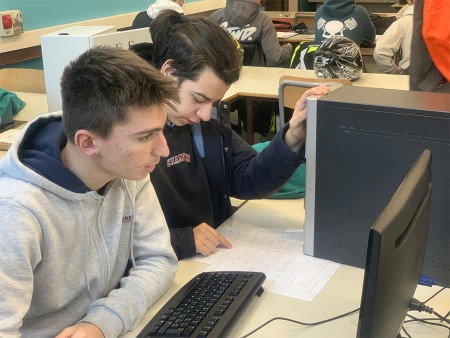  Due studenti lavorano insieme al computer in un'aula, esaminando un foglio con delle annotazioni tecniche. Sullo sfondo si intravedono altri studenti e materiali didattici.