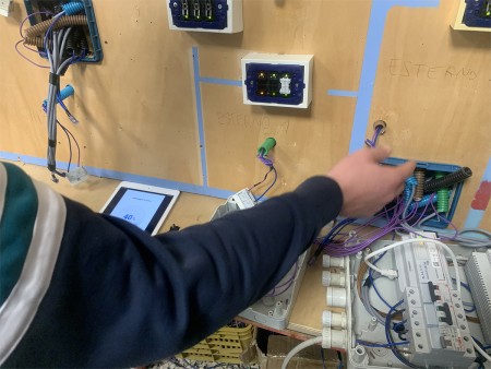 Un dettaglio di un pannello di circuiti elettrici con cablaggio complesso. Una persona è intenta a manipolare i cavi, con un tablet vicino che mostra informazioni o parametri del sistema.