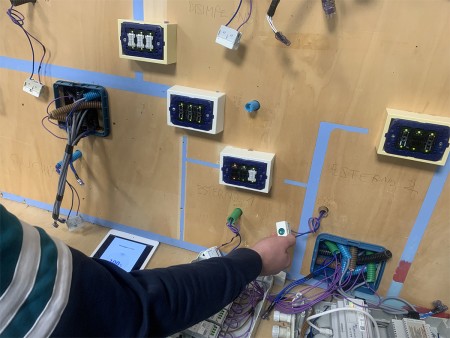 Un pannello elettrico con cablaggi, una persona testa i cavi e un tablet mostra dati del sistema.