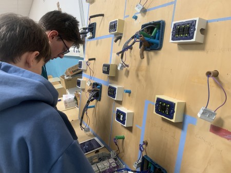 Due studenti lavorano su un pannello elettrico, collegando fili e utilizzando un tablet per seguire le istruzioni.