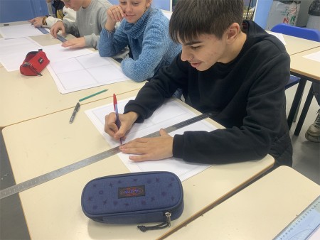 Un ragazzo disegna con righello e penna, mentre una compagna lo osserva sorridendo. Altri studenti lavorano sui loro fogli sullo sfondo.