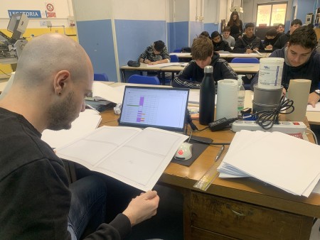 Un insegnante è seduto alla scrivania, mentre esamina e corregge dei fogli di lavoro, utilizzando anche un computer portatile. Sullo sfondo, ci sono studenti concentrati a lavorare sui loro compiti in aula.