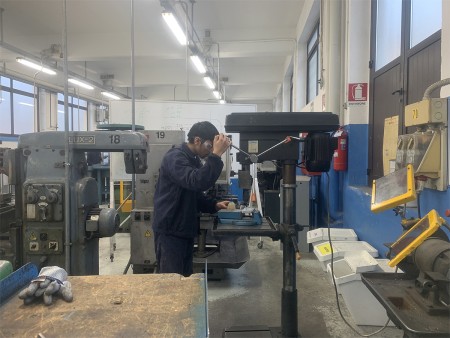 Uno studente sta lavorando su una macchina da officina, indossando occhiali di protezione, in un ambiente industriale. Nella stanza ci sono diversi macchinari e attrezzature per la lavorazione dei metalli, insieme a dispositivi di sicurezza visibili, come un estintore e guanti sul tavolo.
